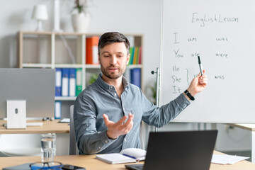 Male english teacher giving remote lesson via laptop, communicating with students on webcam at...