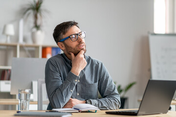 Naklejka premium Thoughtful mature man sitting in office and looking aside, working with laptop, looking for creative ideas