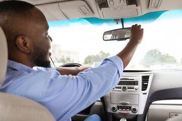 Smiling black guy driving new car in city, adjusting mirror