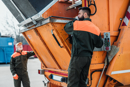 Senior and young garbage men working together on emptying dustbins for trash removal.