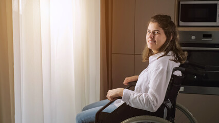 Young disabled woman with organic damage of central nervous system sits in wheelchair looking at camera behind curtain at home, sunlight.
