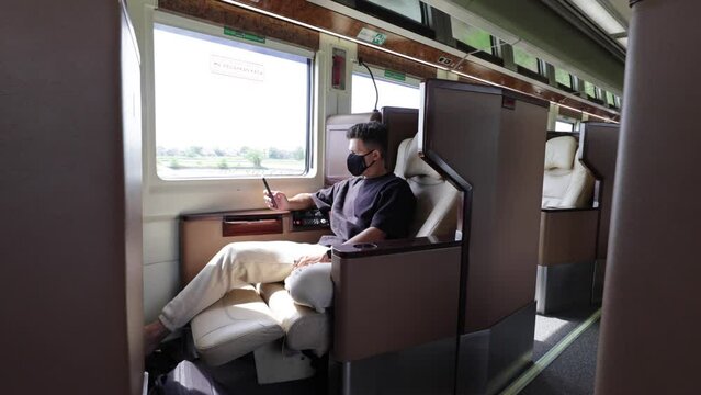 young caucasian male tourist in a mask riding a luxury first class business seat on an Indonesian train looking at a smartphone during the day.