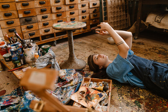 Young woman artist using mobile phone while lying on floor in her studio