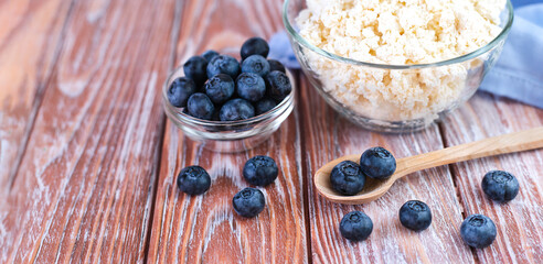 Ripe blueberries in wooden spoon and cottage cheese in a glass bowl on the wooden background. Banner. Close-up. Copy space. Selective focus.