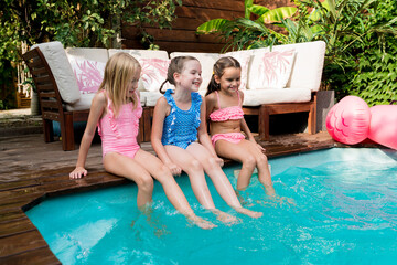 Summertime lifestyle. Girls having fun and relaxing by swimming pool. Kids sitting at poolside and...