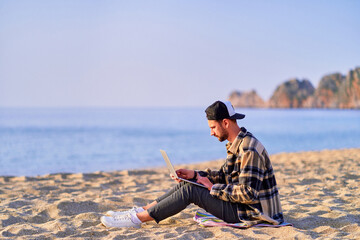 Young happy joyful carefree satisfied millennial freelancer man using laptop on sand beach by the...