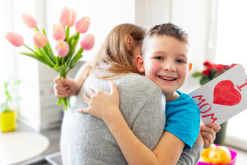 Fototapeta na wymiar Happy Mother's Day! Happy little boy congratulating his mom and giving her flowers tulips and greeting card with red heart drawn