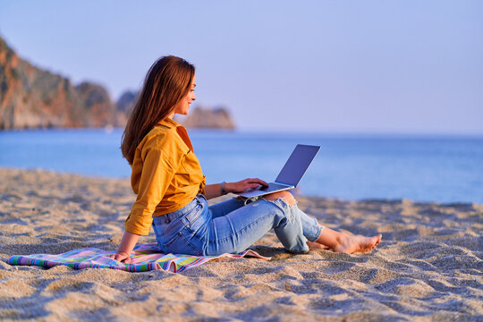 Young happy joyful smiling carefree satisfied millennial freelancer woman using computer and sitting on sand beach by the sea. Dream office remote work concept