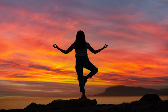 Back view of free calm bliss satisfied woman standing on top rock with yoga position against of idyllic sunset sky by the sea in a happy beautiful inspired moment of her life