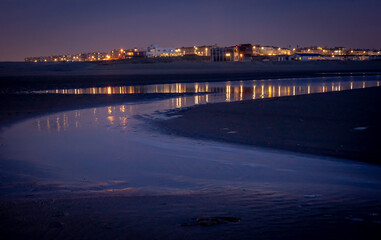 Wieczór nad morzem, plaża, woda wpływająca.  W dali światła miasteczka nadmorskiego.