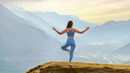 Back view of free calm serene blissful satisfied fitness woman doing yoga exercise on top rock against idyllic morning mountain view. Mental mind care and healthy habits