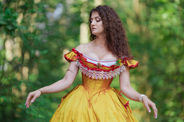 A young woman in an ancient medieval yellow orange dress walks in a green park on a summer day