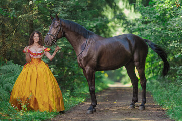 A young woman in a vintage yellow dress walks with a brown horse in a green park on a summer day