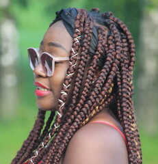 Portrait of a young African woman with dreadlocked pigtails on her head. A young adult girl from Africa in sunglasses in nature.