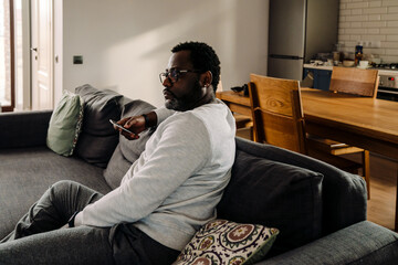 Black man wearing eyeglasses watching tv while resting on sofa