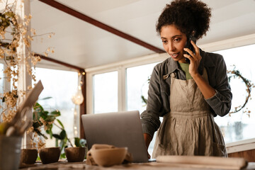 Young black ceramist woman using cellphone and laptop in her studio