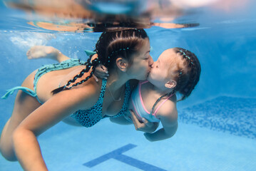Mother kissing baby daughter learn to swim on swimming lesson, doing physical exercises, dive underwater with in pool. Healthy lifestyle, active parents. Pregnant woman swims underwater.