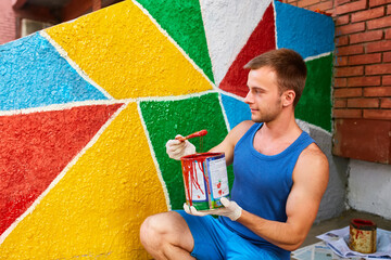 The guy paints the wall with a brush in bright colors, holds a can of paint in his hand