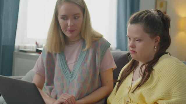 Young woman explaining how to use laptop to girl with Down syndrome while sitting together on couch at home