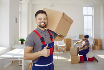 Smiling male worker of moving and delivery company holding cardboard box showing thumbs up. Loader in overalls posing against background of colleague who packs cardboard boxes. Moving service concept.