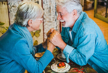 Smiling attractive old senior couple hands in hands looking in the eyes at coffee shop. Cappuccino and fruit cake on the table