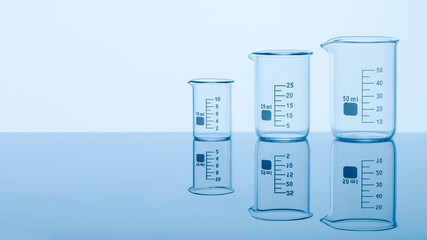 Three empty measuring beakers sitting on a mirror blue surface, glass lab containers standing in gradual order on a table, transparent light through laboratory flasks, precise comparing measurements 