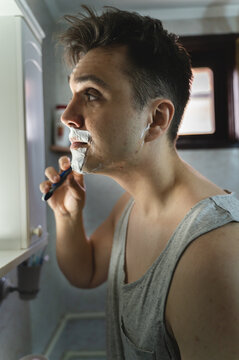 A young man is shaving in bathroom while looking at mirror during the day