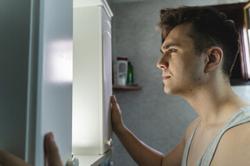 A young man is shaving in bathroom while looking at mirror during the day