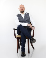 Portrait of serious middle-aged businessman in grey vest, blue jeans, white shirt, sitting on chair with legs crossed.