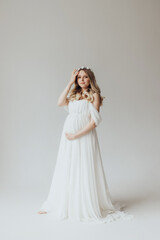 Pregnant blonde girl in a white dress in the studio with a wreath on her head