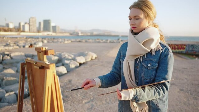 Creative hobby. Young inspired woman artist drawing picture of seaside, standing alone on bank with easel, tracking shot