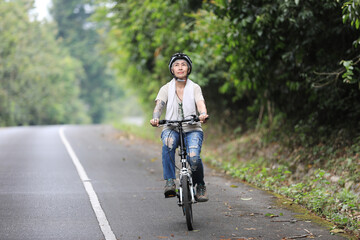 Healthy elderly woman riding bicycle with stylish happy face. Exercise for health, wear a helmet on the green road, there are only trees on both sides, enjoy watching nature in the park in retirement