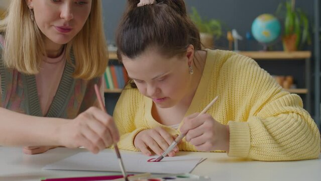 Girl with Down syndrome and female teacher painting picture together at desk during art class at home