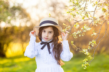 Portrait of beautiful little girl in hat and white dress , standing under flowering trees, in spring