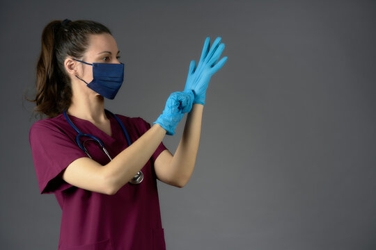 Nurse In Purple Uniform Mask And Stethoscope Putting On Latex Medical Gloves On Gray Background