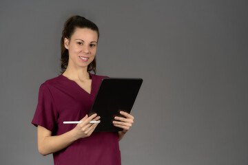 smiling female nurse in purple uniform looking at camera with digital tablet report on gray...