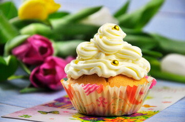 A festive cupcake with white cream and flowers in the background.