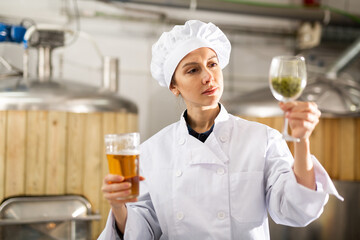 Successful professional female brewer supervising production process in brewery, holding glass of...
