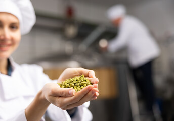 Unrecognizable woman brewer in white coat holding bunch of hops pellets.