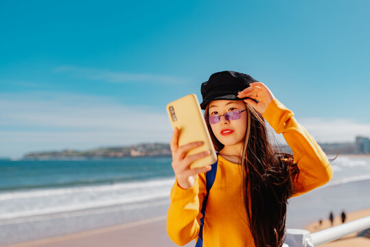 young asian woman takes a picture of herself with her mobile phone while sightseeing on a city beach. chinese girl taking a selfie