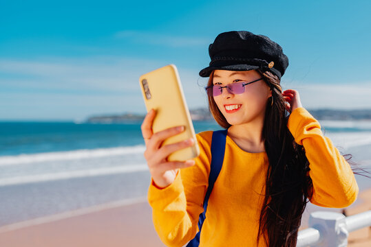 young chinese woman in beret, yellow sweater and purple sunglasses takes pictures of herself posing on the street. Asian girl takes a selfie with her smartphone.