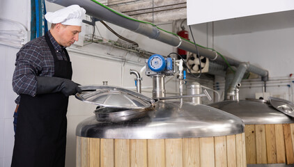 Focused skilled brewmaster opening lid of fermenter while checking process of craft beer production...