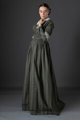 A Victorian working class woman wearing a checked bodice and skirt and standing alone against a studio backdrop