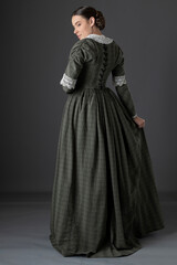 A Victorian working class woman wearing a checked bodice and skirt and standing alone against a...
