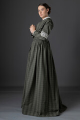 A Victorian working class woman wearing a checked bodice and skirt and standing alone against a studio backdrop