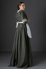 Fototapeta premium A Victorian woman wearing a dark green checked bodice and skirt and standing alone against a studio backdrop