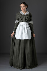 A Victorian woman wearing a dark green checked bodice and skirt and standing alone against a studio...