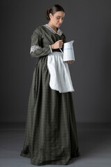 A Victorian working class woman wearing a dark green checked bodice and skirt and standing alone against a studio backdrop