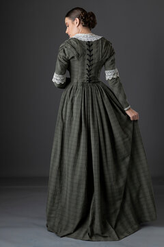 A Victorian working class woman wearing a checked bodice and skirt and standing alone against a studio backdrop