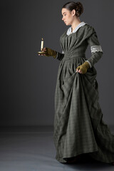 A Victorian working class woman wearing a dark green checked bodice and skirt and carrying a candle...